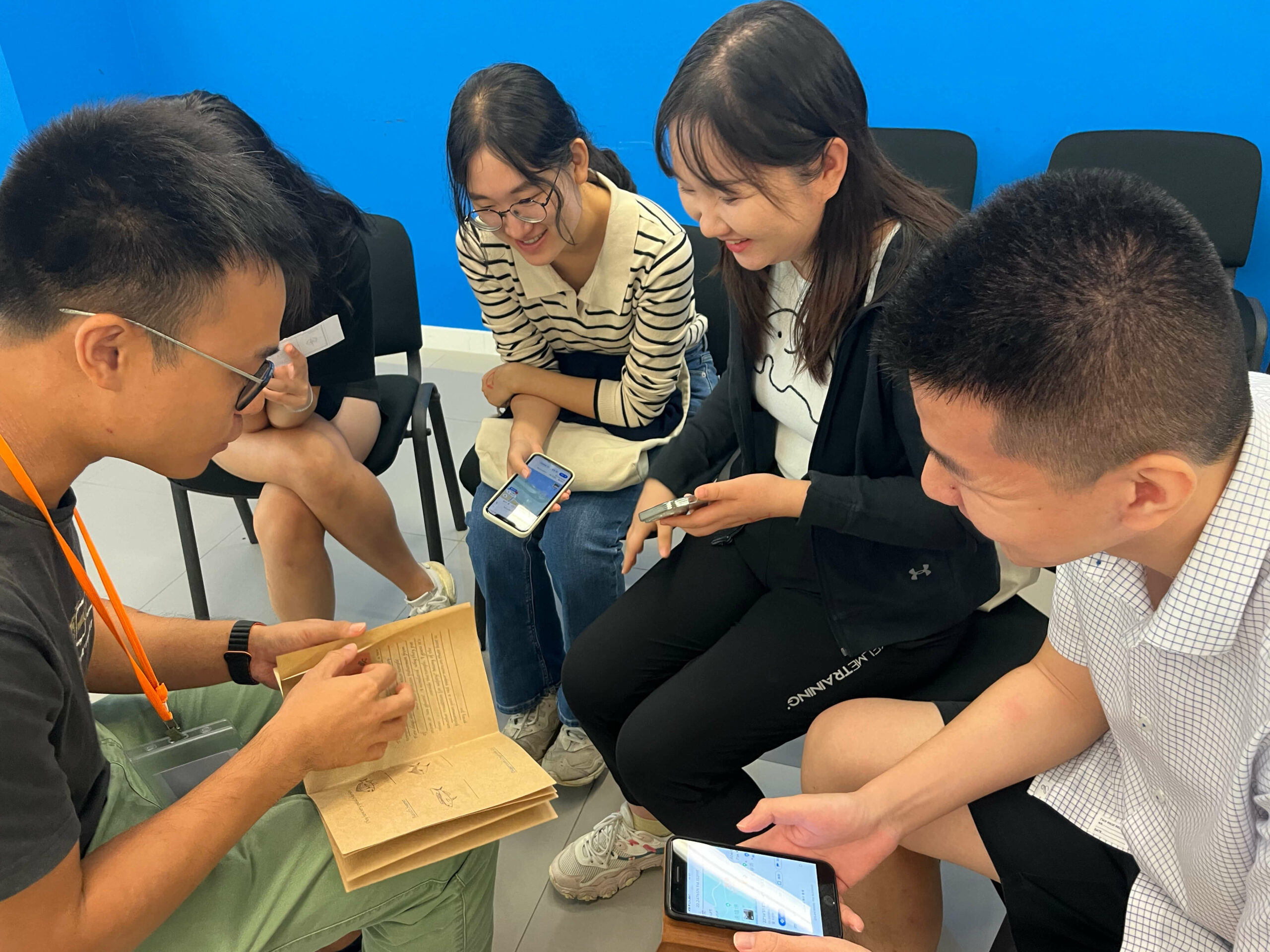 Four participants are seated together with the instructor, engaging with books and a smartphone.