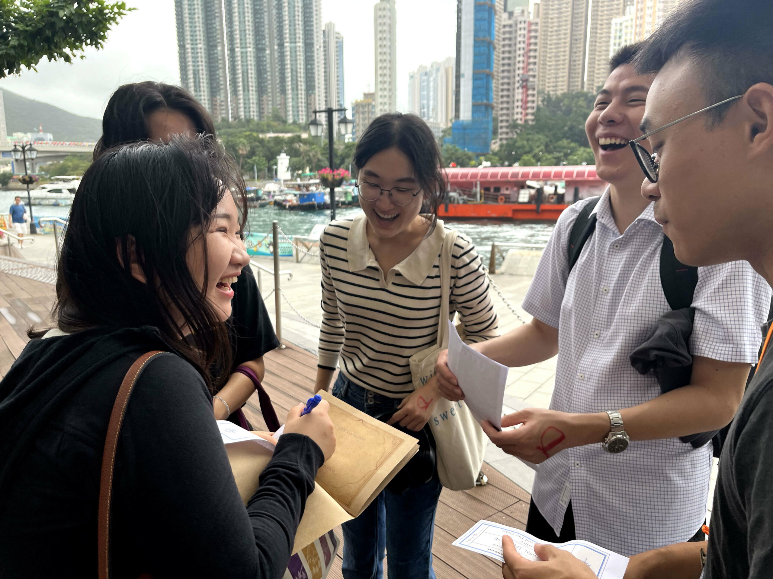 A group of people stands near a waterfront in an urban setting with high-rise buildings. One person holds an envelope and writes on the map, looking for the next checkpoint of the game.