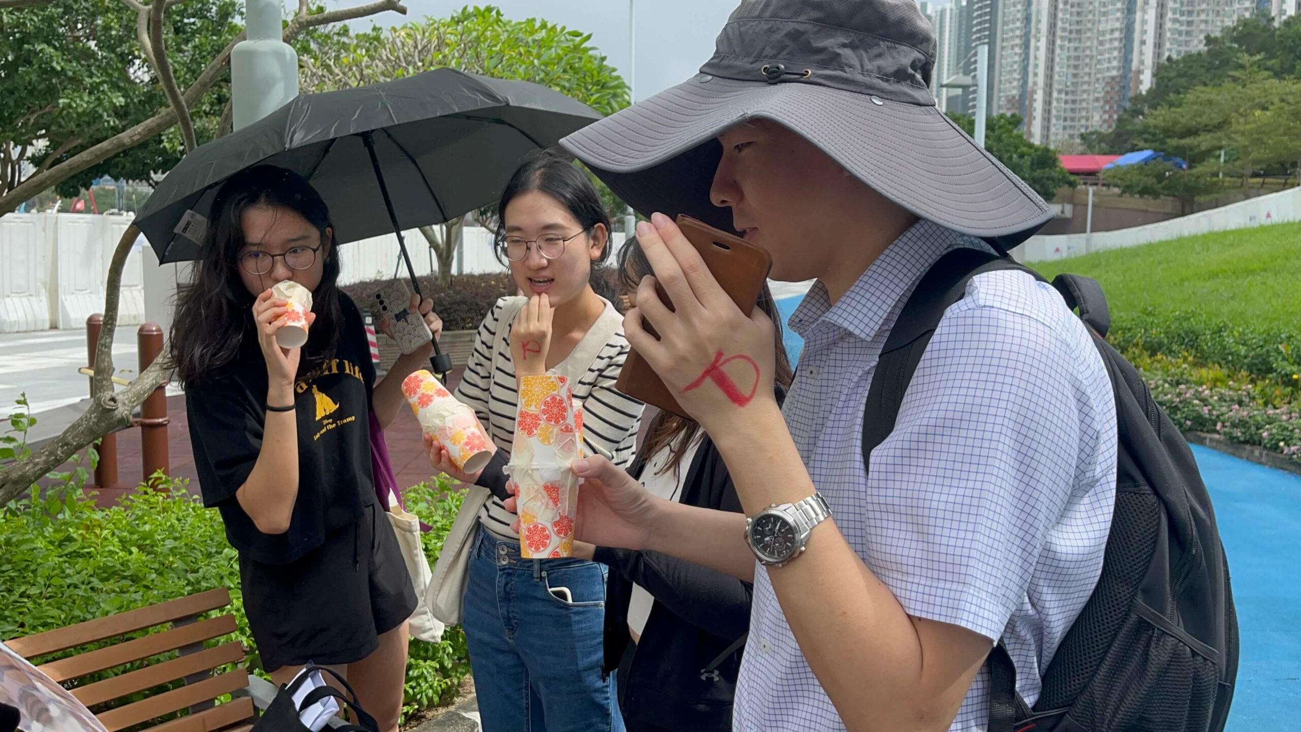 A group of people standing outdoors, holding colorful drinks in plastic cups. One person is holding an umbrella, and another has a red mark on the hand.