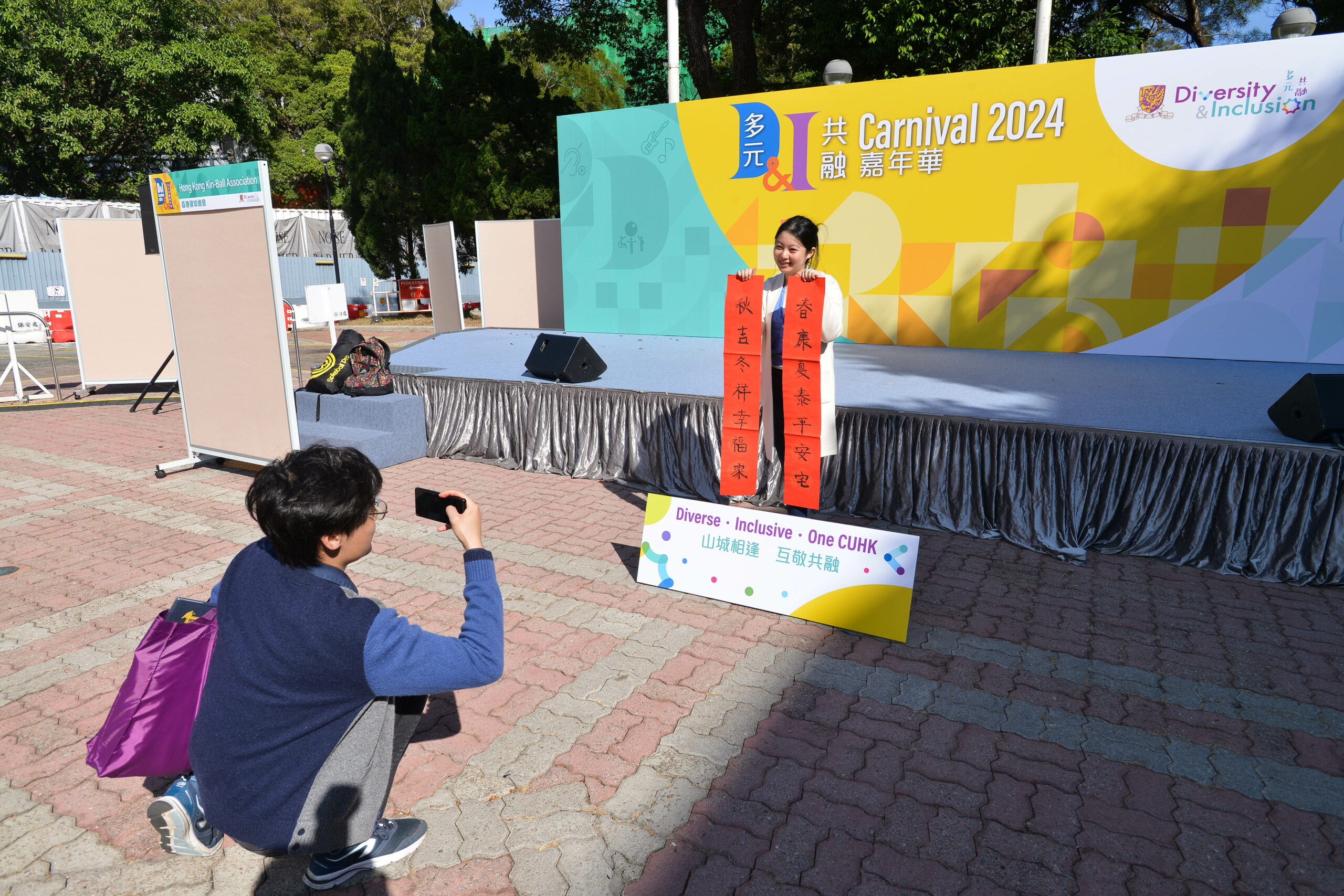 A person holds two fai chun. Another person is kneeling in front of the stage, taking a photo with a smartphone.