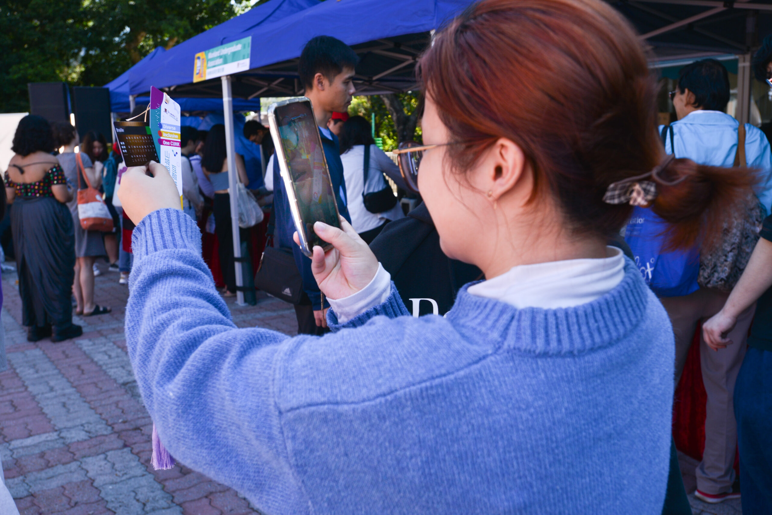 A student taking a photo with a smartphone at the Carnival, holding a bookmark made at the workshop. Crowd and a booth with a blue canopy in the background