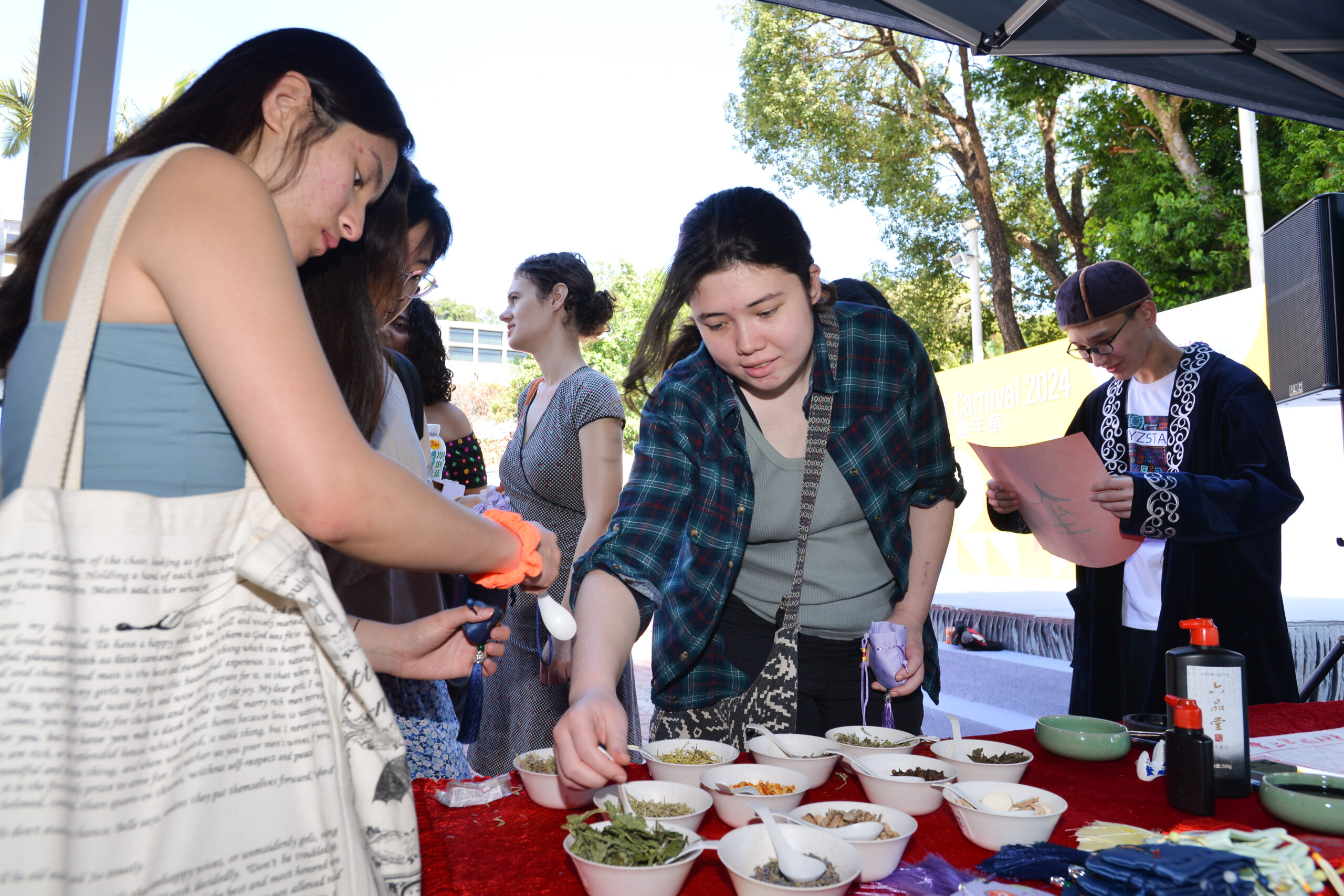 Group of people at D&I Carnival 2024, gathered around a table with bowls containing dried herbs or spices, in an outdoor setting