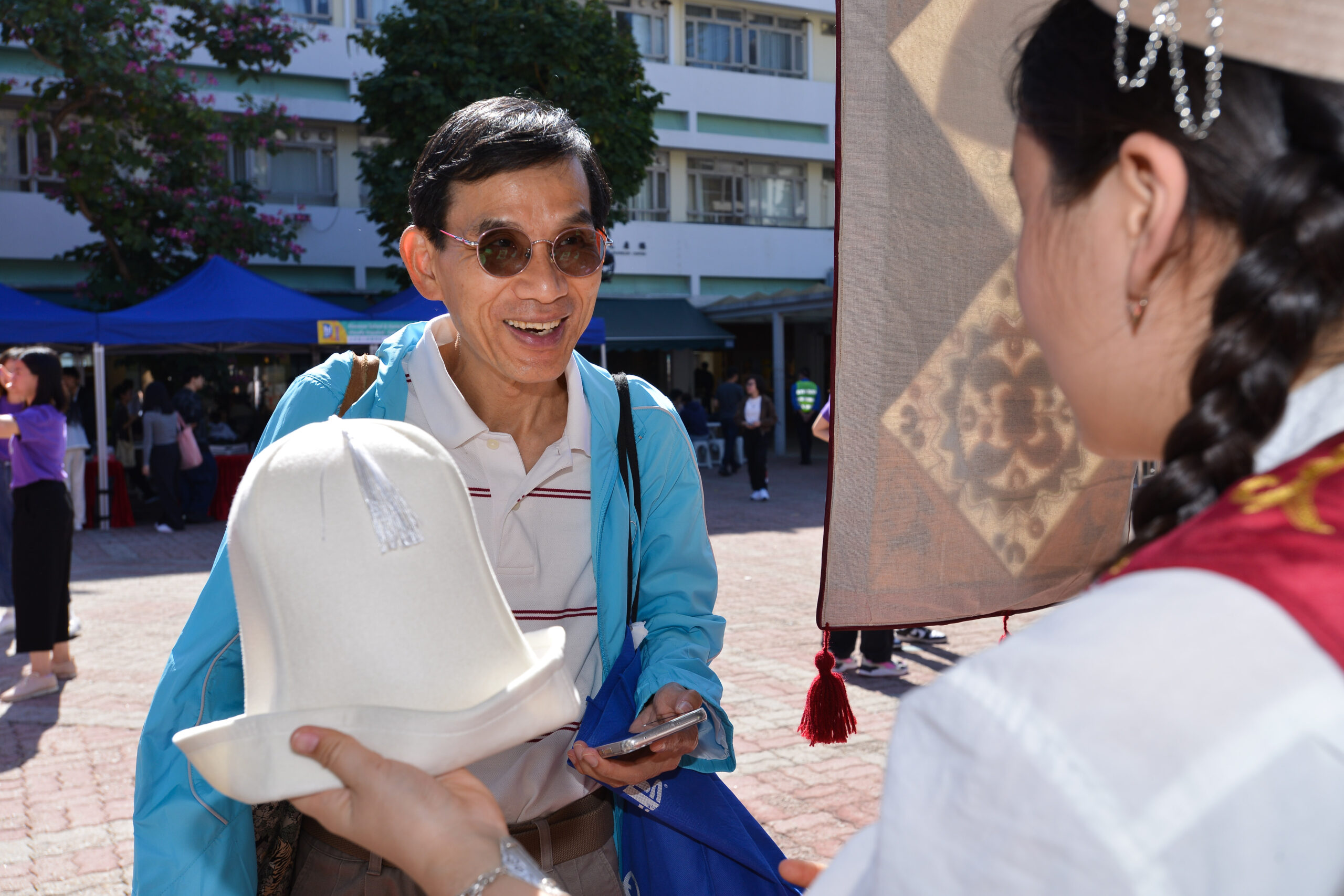 A participant holding a white hat with a tassel in front of another person wearing a light blue jacket, outdoors near a building with a blue tent.