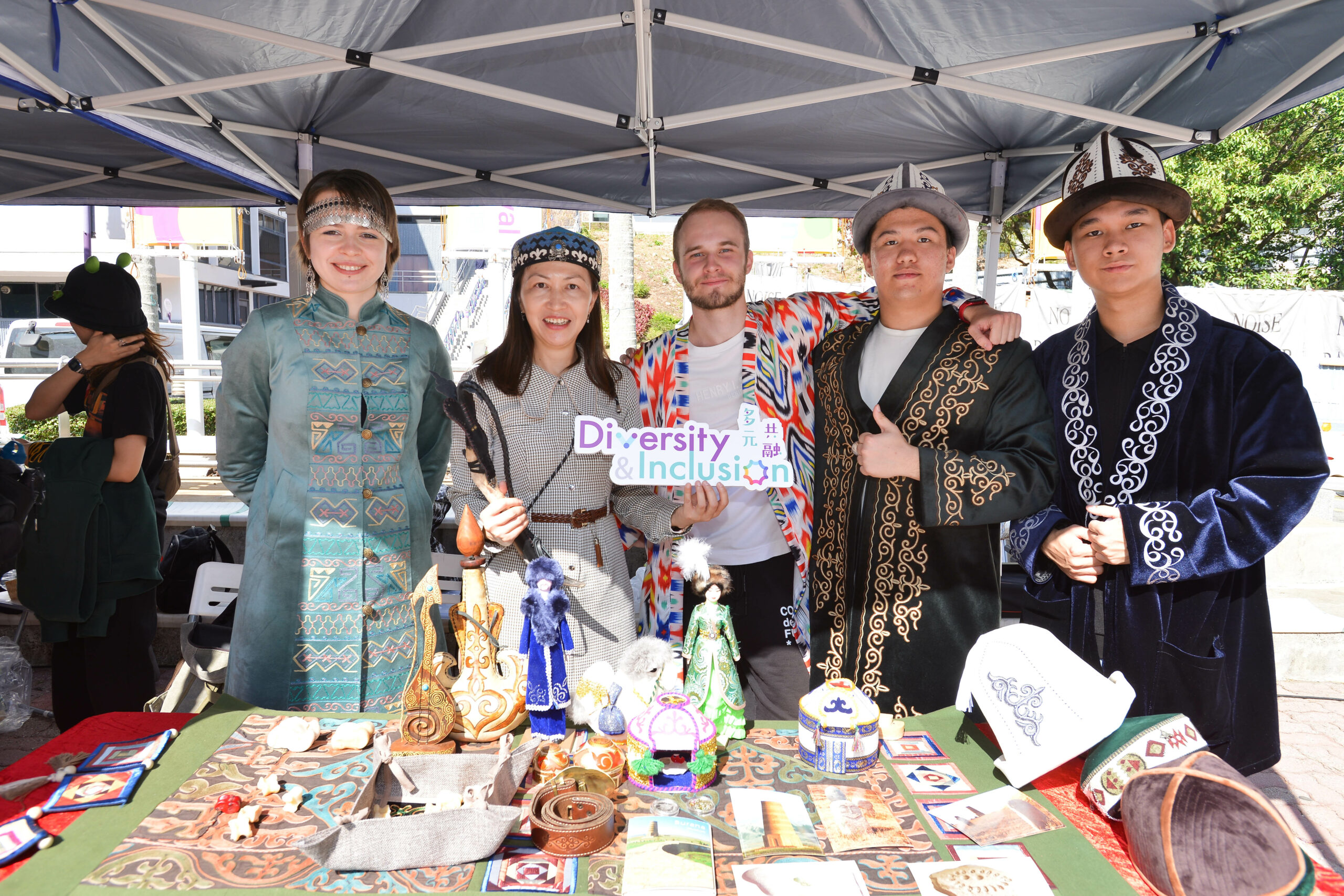 Participants at D&I Carnival 2024, standing under a canopy in traditional attire, showcasing cultural items and a 'Diversity & Inclusion' sign.