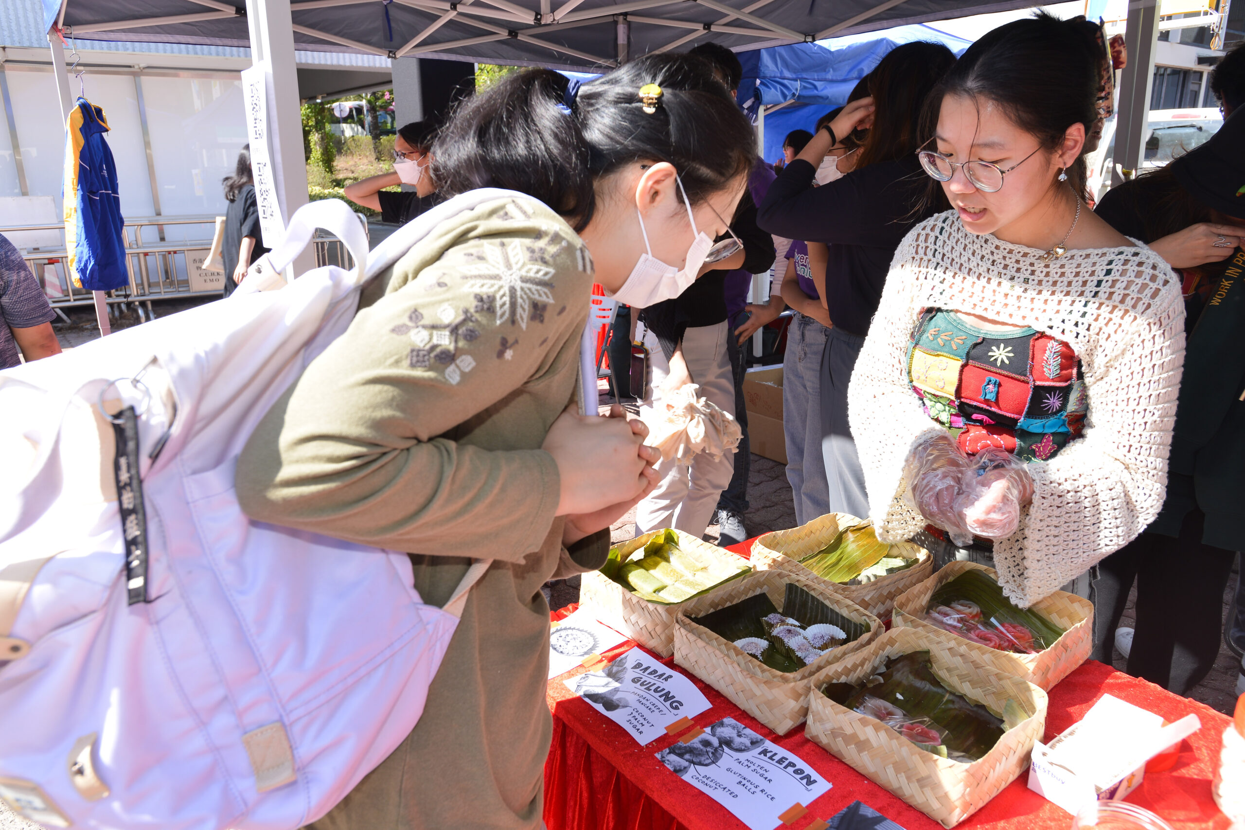 Participant at a D&I Carnival 2024 stall with 'DADAR GULUNG' and 'KLEPON' food items displayed in woven baskets with labels.