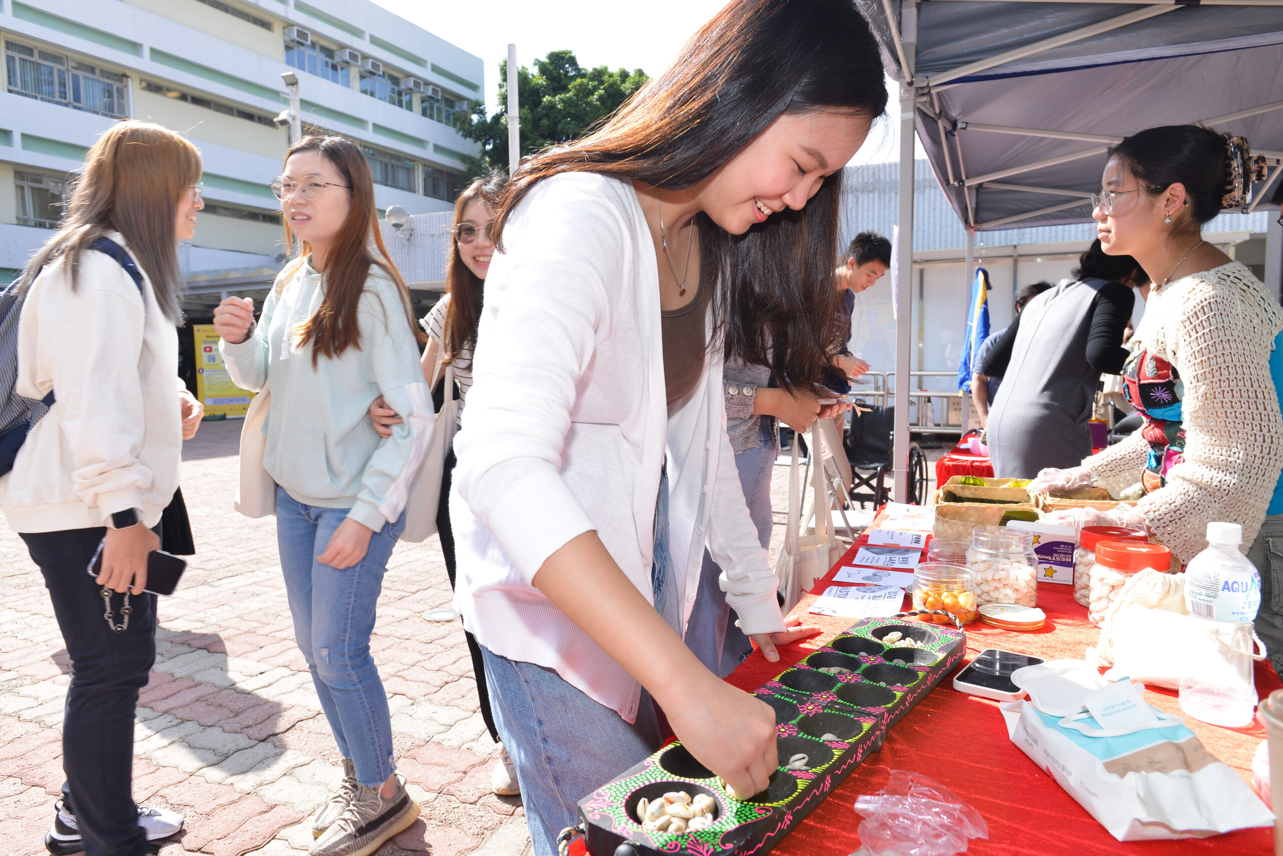 Participants at D&I Carnival 2024 gathered under a canopy, engaging with items on a table that include colorful trays, jars, and papers.