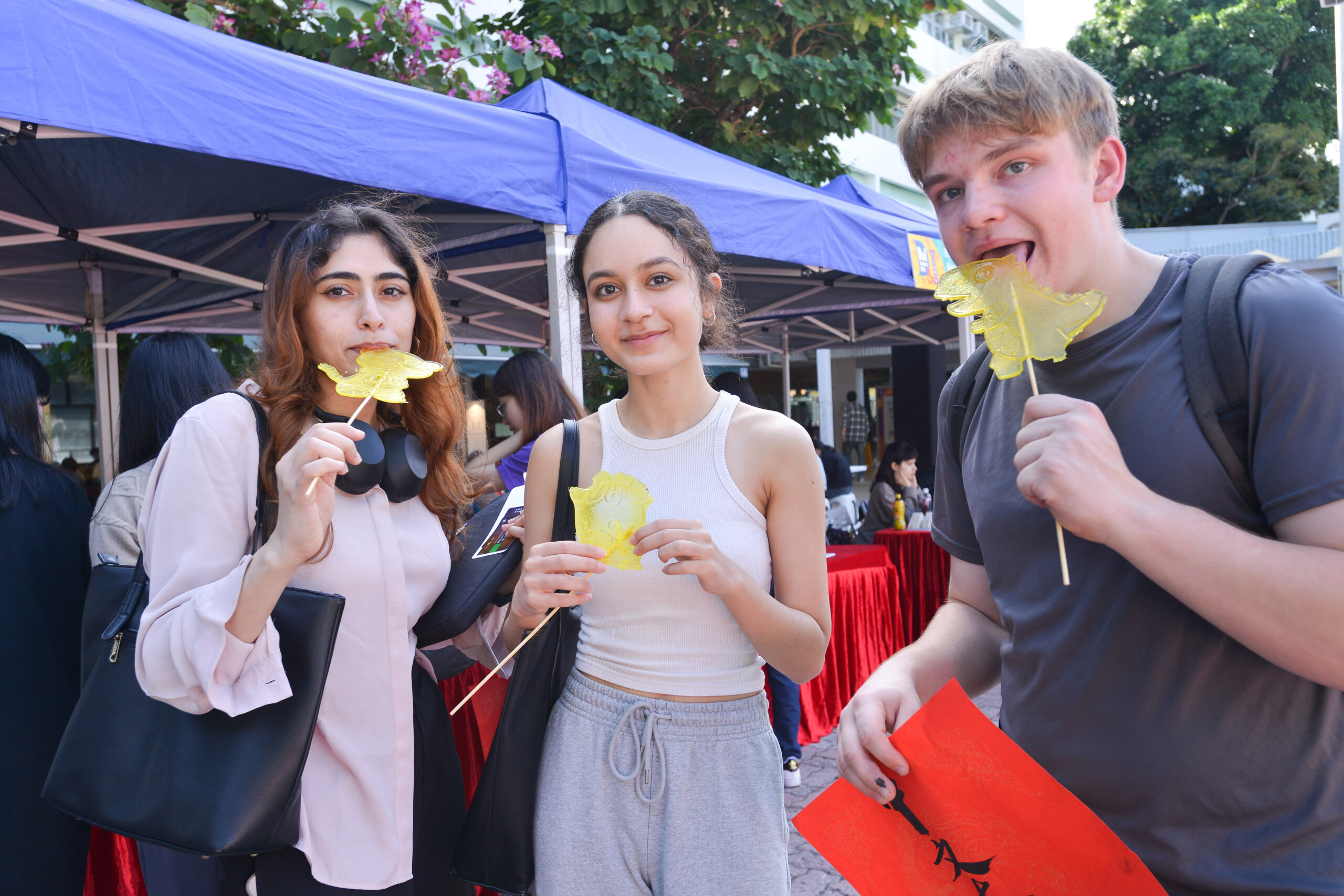 Participants at D&I Carnival 2024 standing in front of a booth with a blue canopy, holding yellow leaf-shaped snacks and a fai chun..