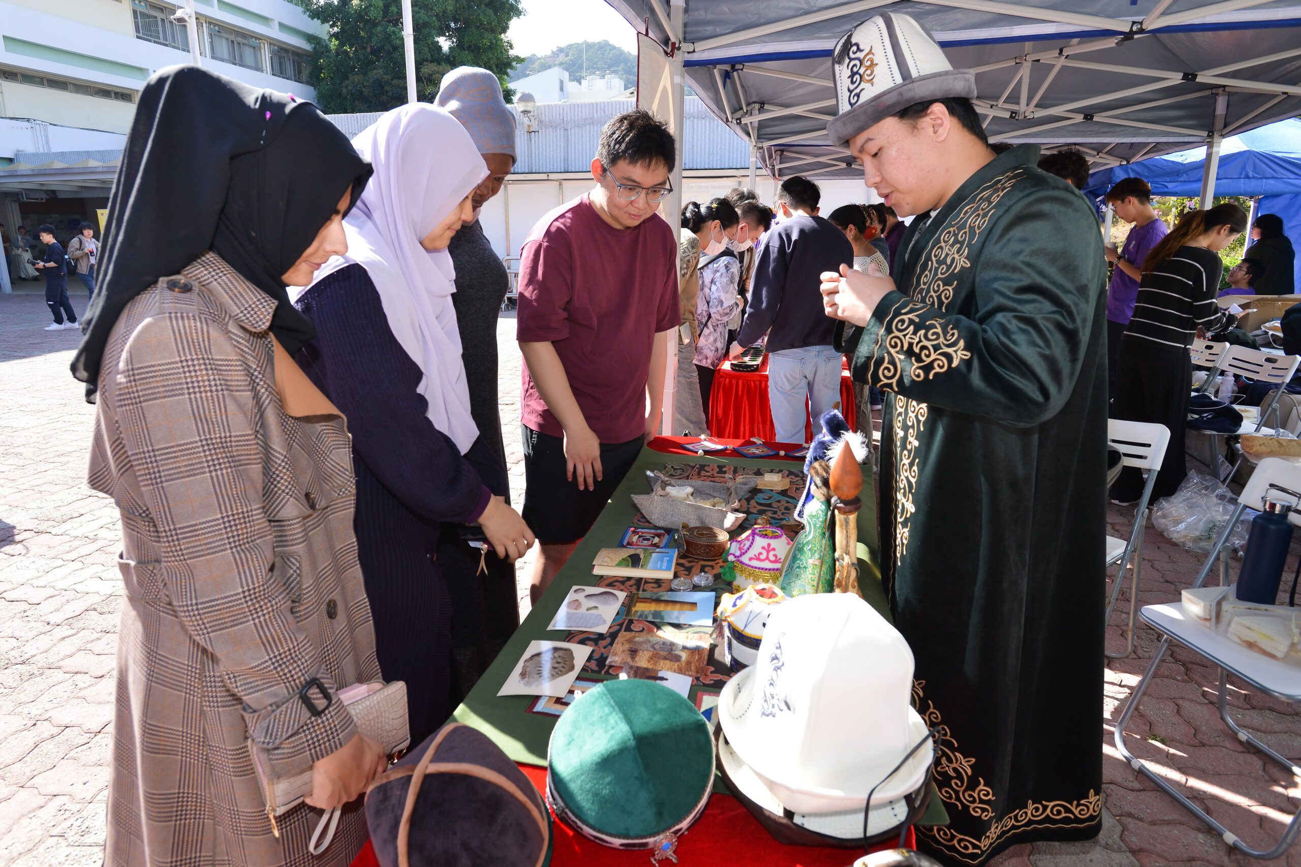 Group of people at the event booth under a tent, displaying various traditional hats, books, and cultural artifacts on a green cloth-covered table.