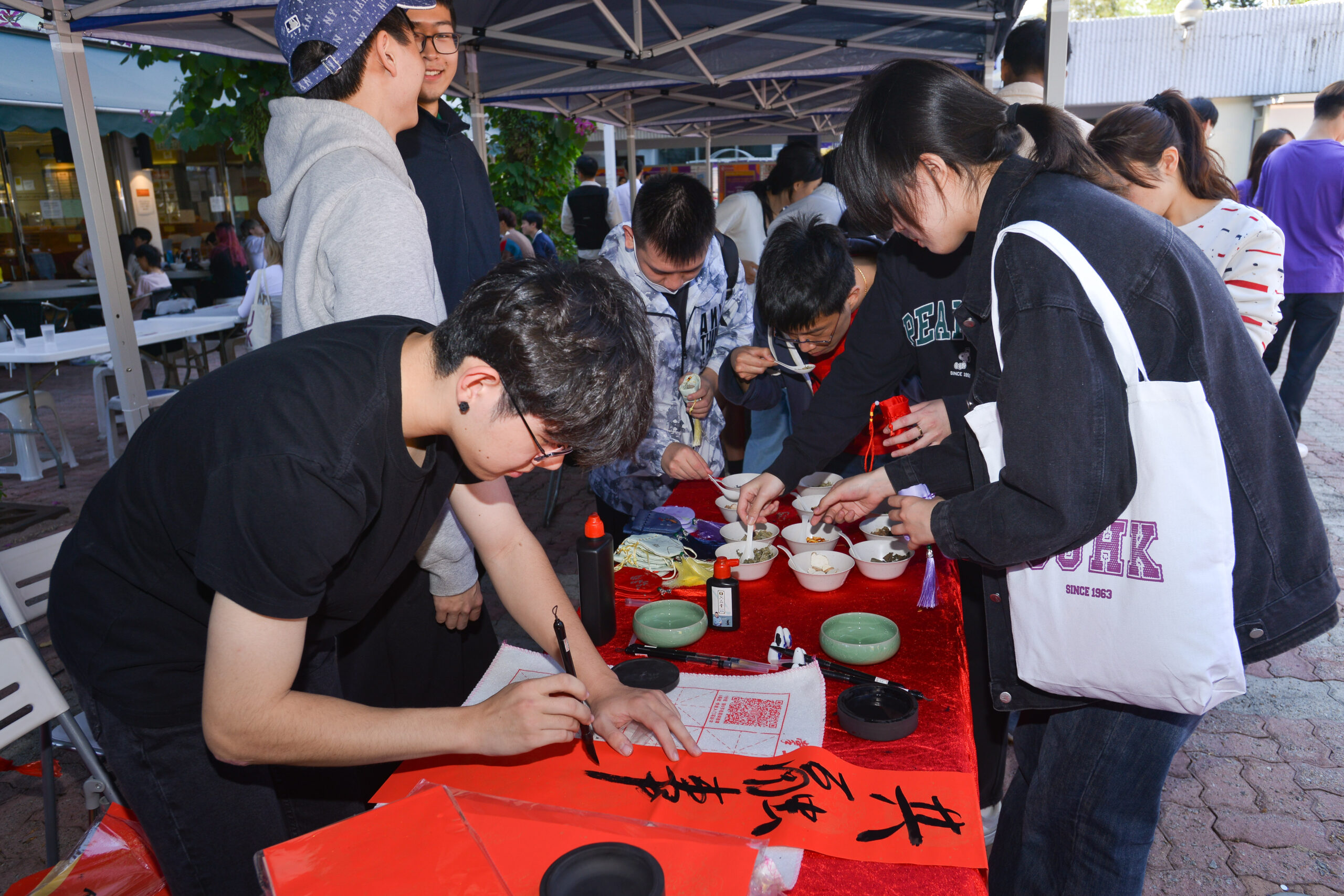 Participants at D&I Carnival 2024 writing fai chuns under a canopy, with various calligraphy supplies on the table.