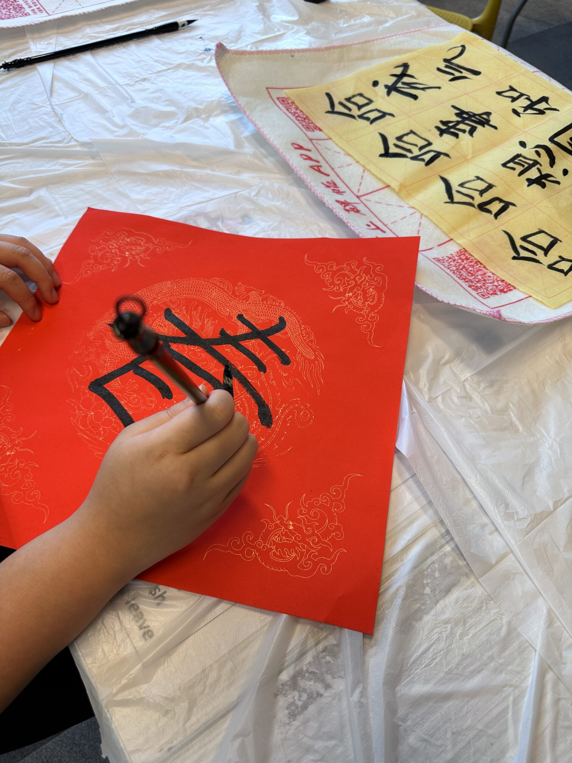 A person practices Chinese calligraphy at a Fai Chun workshop, writing characters on a red paper with intricate dragon designs using a brush.