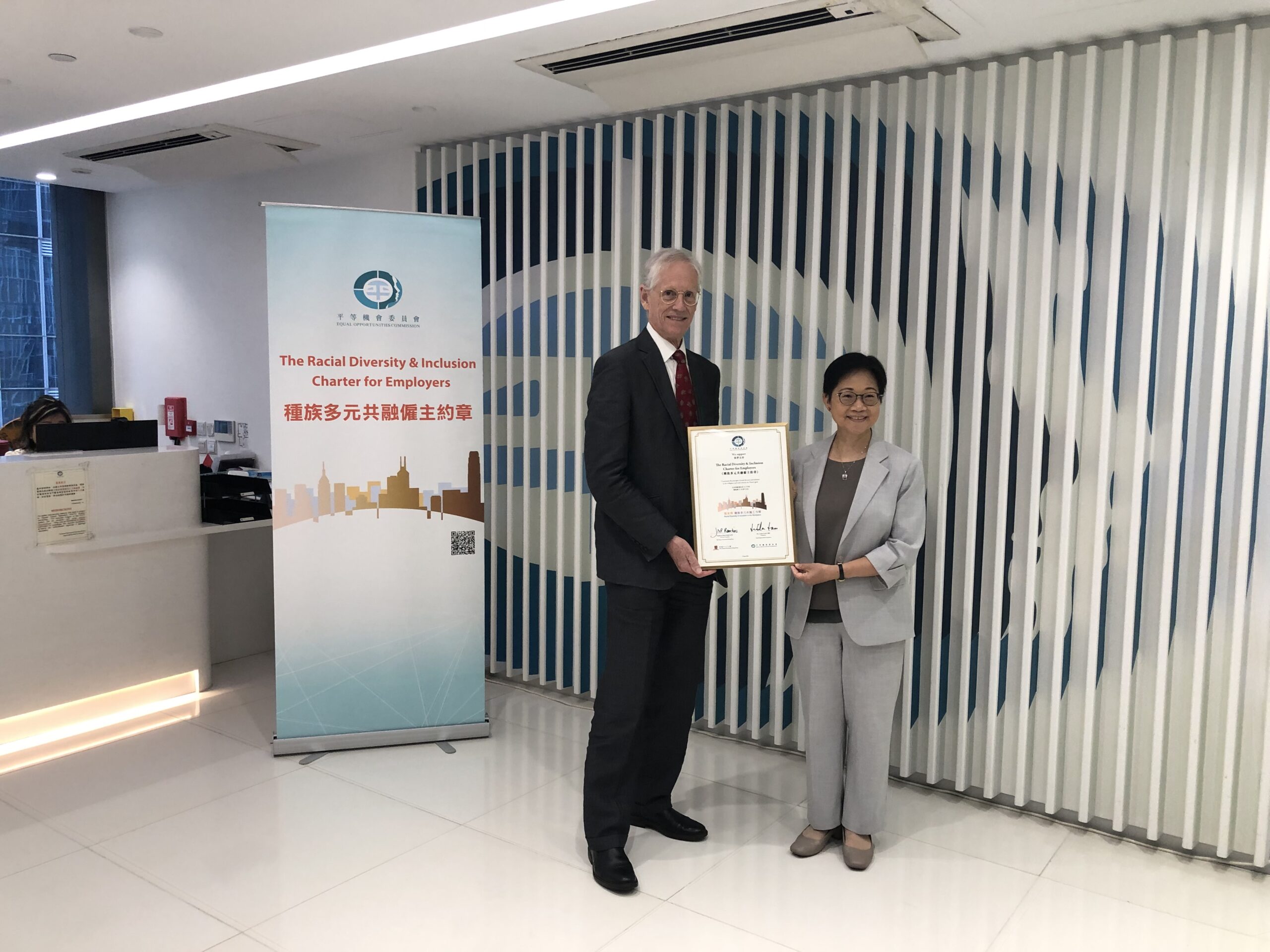 Ms. Linda Lam and Prof. Nick Rawlins holding a framed certificate stand in an office with a "Racial Diversity & Inclusion Charter for Employers" banner in English and Chinese behind them.