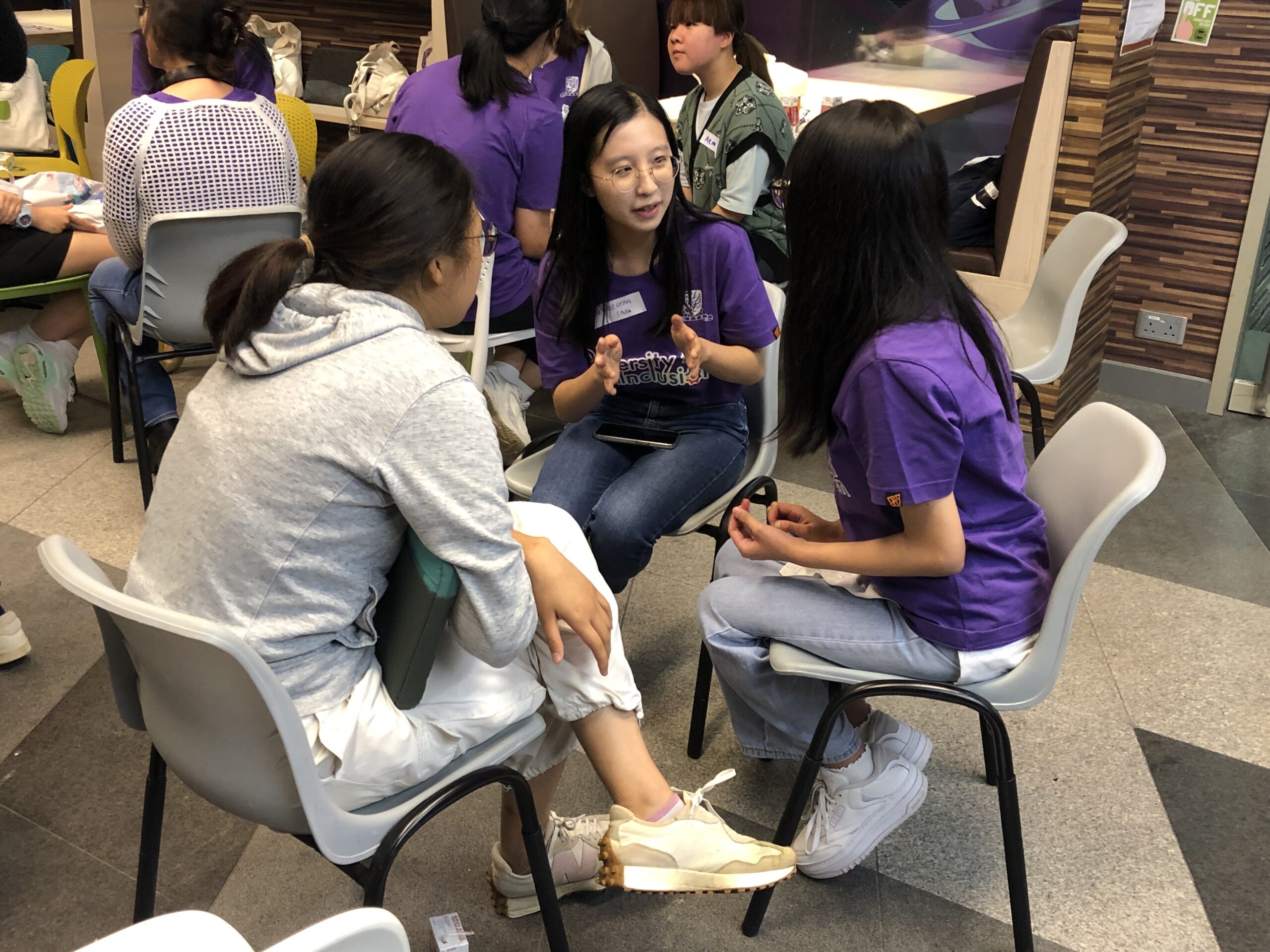 A group of ambassadors sits in a circle, engaged in a discussion. Some wear purple D&I shirts. The room has wooden panel walls and posters.