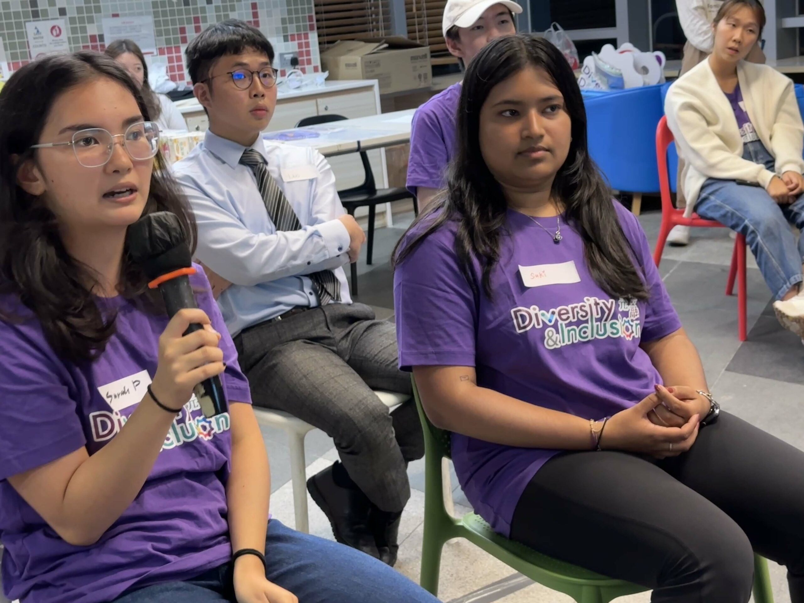 A group of D&I ambassadors, seated in a room, with some wearing purple "Diversity & Inclusion" t-shirts. One holds a microphone, suggesting participation in a discussion.