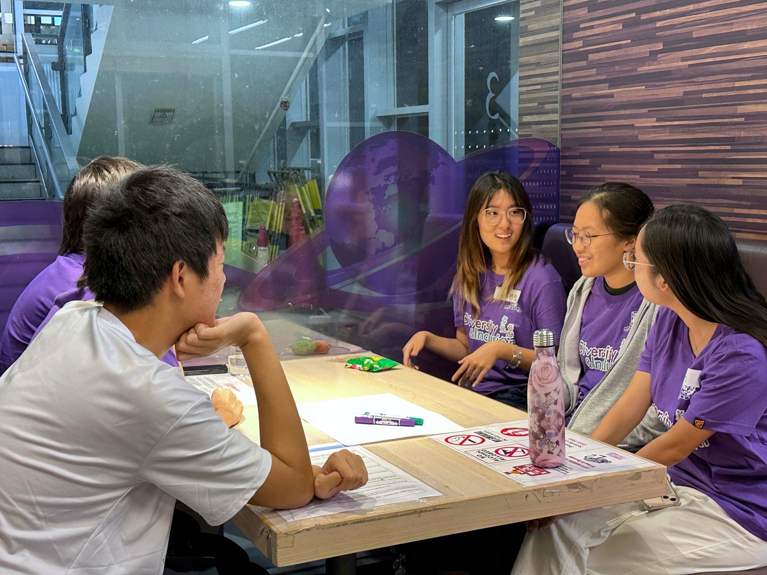 A group of D&I ambassadors sits around a table during a workshop. They are actively engaged in a discussion with papers, markers, and drinks on the table.